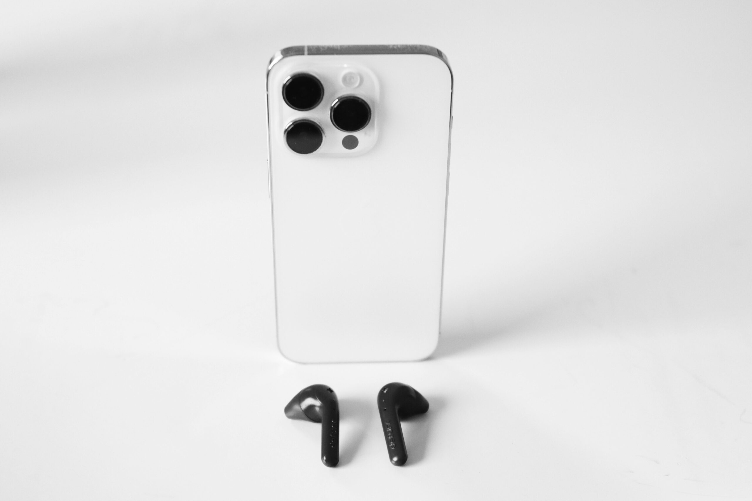 A minimalist still life of a smartphone with black AirPods on a white surface, showcasing modern design.