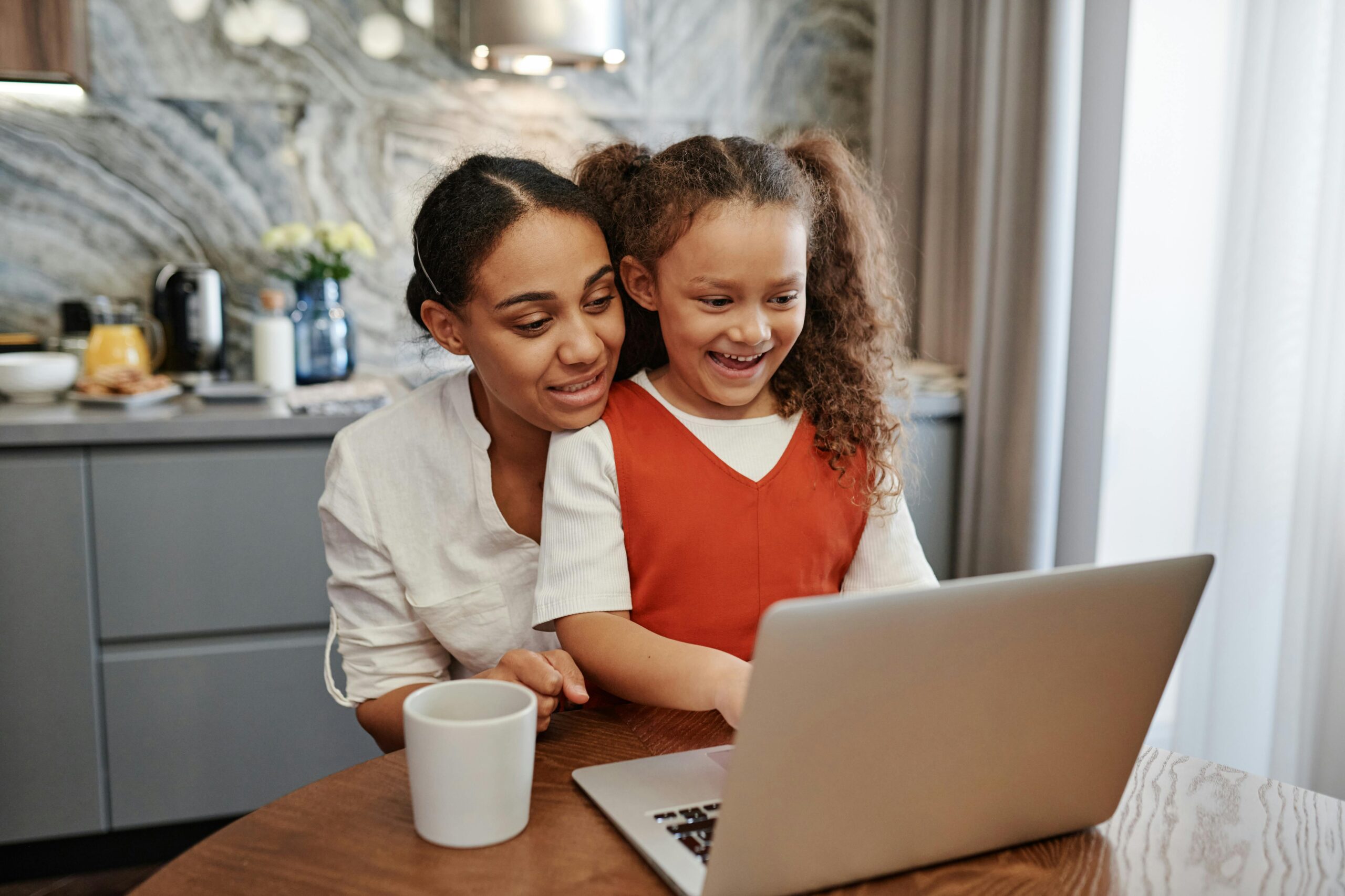 A mother and daughter smiling while using a laptop together in the kitchen.